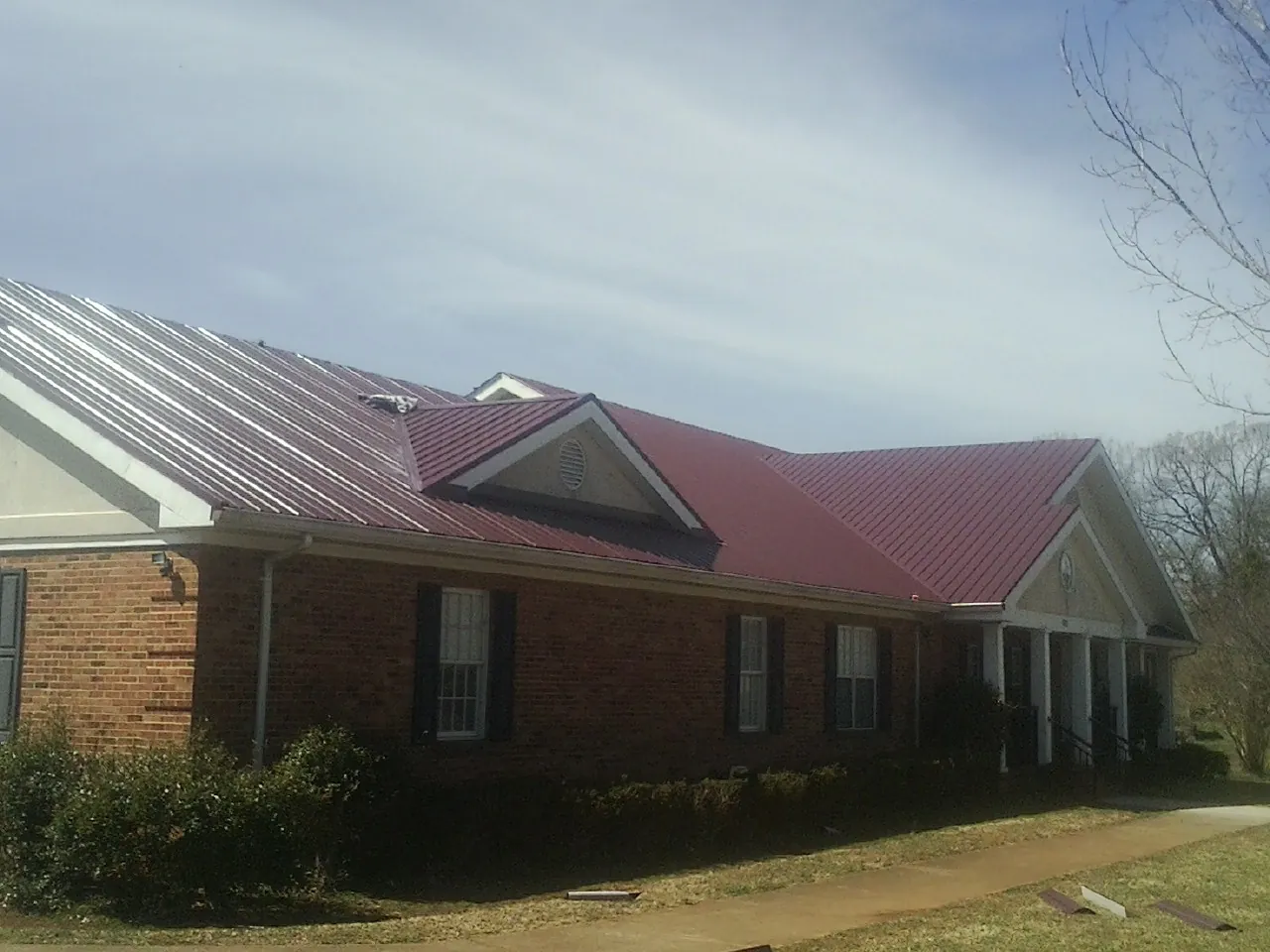 Red metal roof with gabled entries on a long brick building, from a Hail Damage Roof Repair job in Myton
