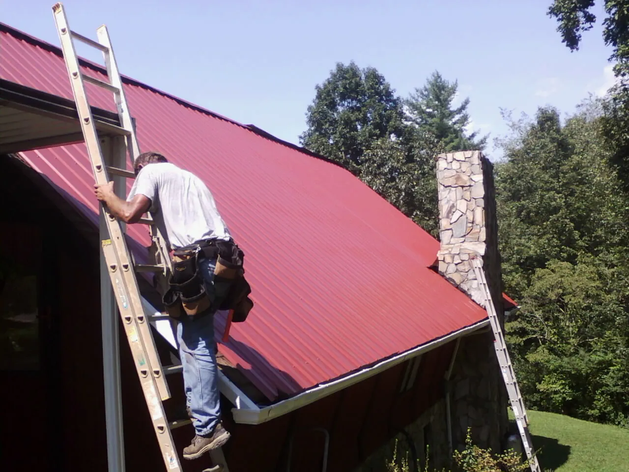 Roofer on a ladder beside a red metal roof and stone chimney — Soffit & Fascia Repair by Roofing Brothers in Myton
